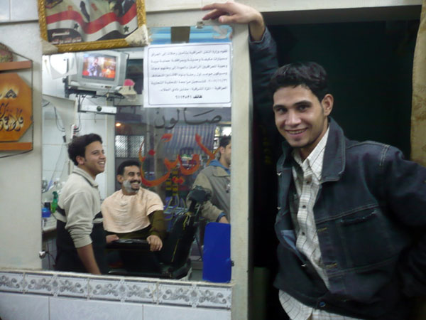 A notice in an Iraqi-owned barber shop in Damascus advertises free trips for returnees. The customer being shaved has no intention of returning. The barber and his friend say, "Perhaps, but not yet."