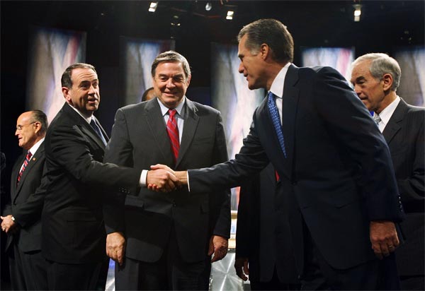 Mike Huckabee and Mitt Romney shake hands before a debate, while Duncan Hunter looks on.